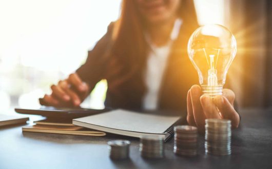 Businesswoman holding a lightbulb with coins stack on table, saving energy and money concept
